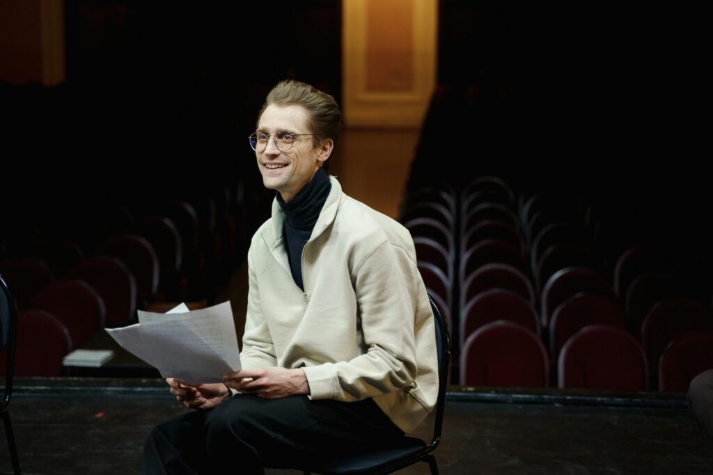 Smiling actor rehearsing script alone in empty theater auditorium, seated on stage.